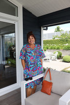 Woman in a colorful dress standing on a balcony with a view of greenery and a building.