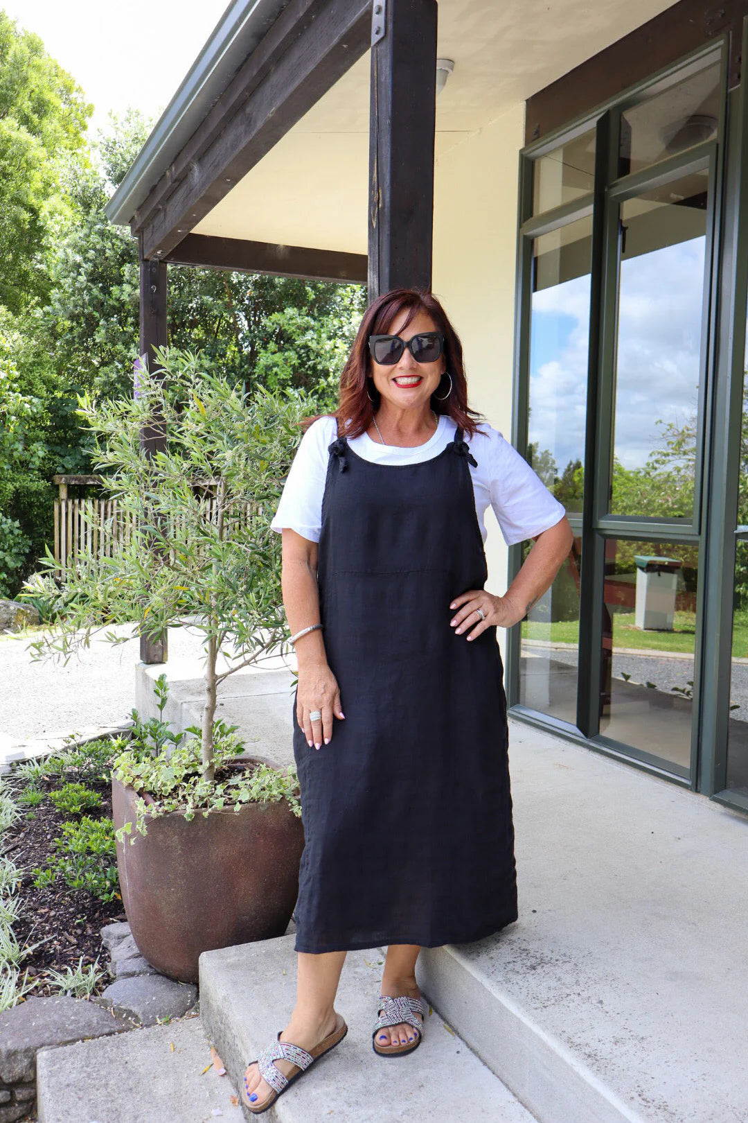 Woman wearing a black dress and sunglasses standing outside a building with greenery.